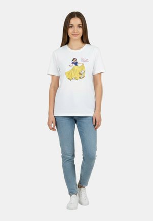 Giovane donna in piedi che indossa una t-shirt bianca con grafica colorata di Biancaneve e jeans azzurri su sfondo neutro.