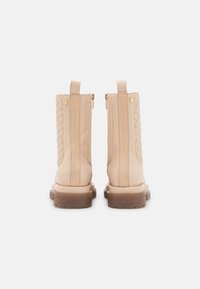 Botines beige hasta el tobillo con lados acolchados, acentos dorados, cremalleras traseras y una suela de goma marrón gruesa. Acabado en cuero suave con detalles de marca.