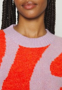 Pull en tricot avec un motif violet et orange. Texture douce, encolure ronde, et couleurs contrastées créent un design visuel audacieux.