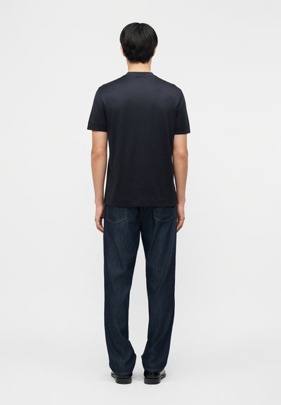 Άνδρας που στέκεται με την πλάτη, φοράει απλό μαύρο κοντομάνικο πουκάμισο, σκούρο μπλε τζιν και μαύρα παπούτσια σε απλό φωτεινό φόντο.