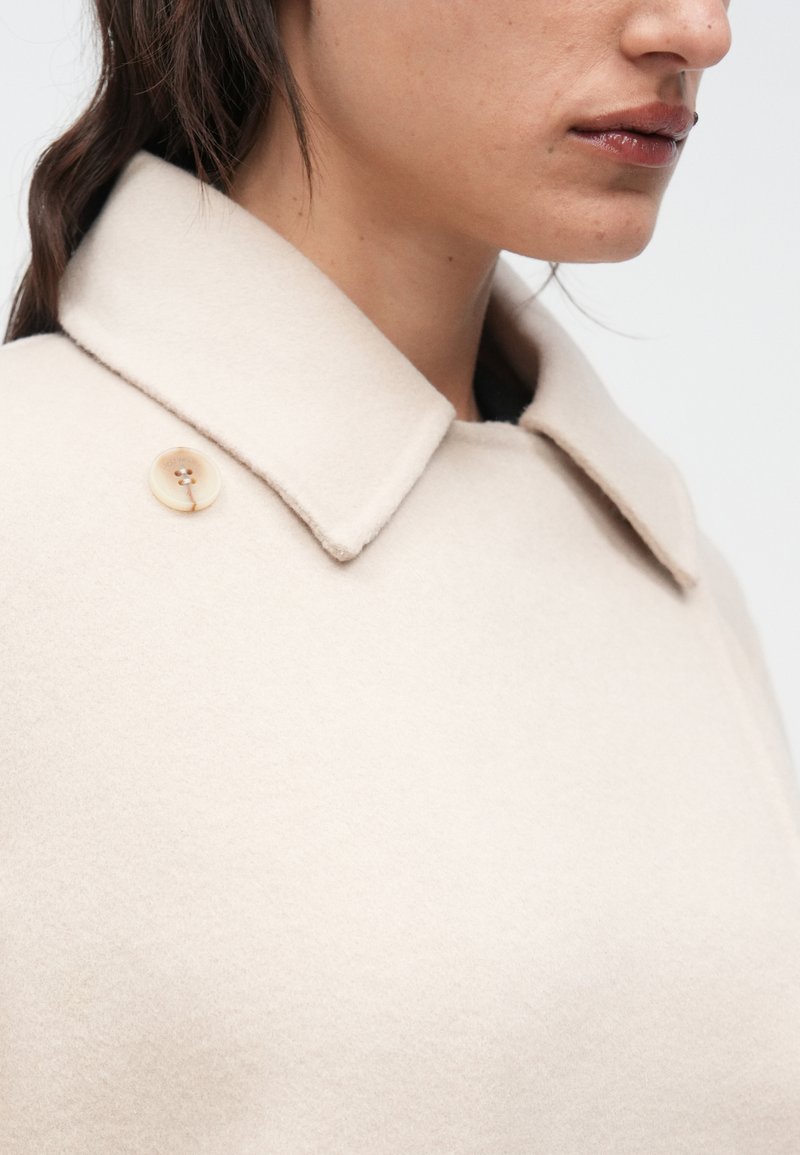 Beige wool coat with a structured collar and a single button detail. Soft texture, minimalist design, and subtle stitching accents.