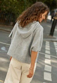 Világosszürke kötött kapucnis pulóver bordázott szegéllyel és rövid ujjakkal, bézs színű, széles szárú nadrággal párosítva, utcai háttér előtt.