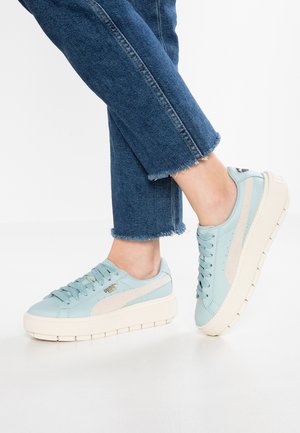 Ben iført blå jeans og lyseblå Puma-sneakers med tykke hvide såler, stående på en hvid overflade.