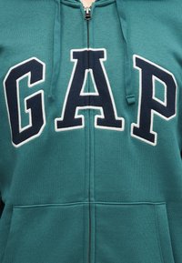 Sweat à capuche zippé couleur sarcelle avec les grandes lettres "GAP" en bleu marine et blanc en contour sur la poitrine et les poches avant.