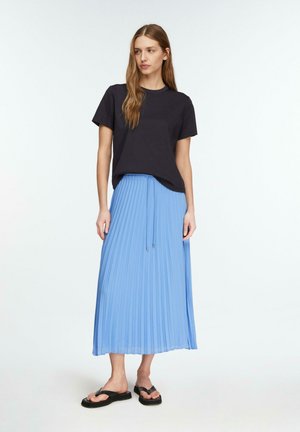 Frau trägt ein schwarzes kurzärmeliges Shirt, hellblauen plissierten Midirock mit Kordelzug an der Taille und schwarze Pantoletten, steht vor weißem Hintergrund.