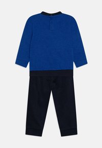 Maglietta blu a maniche lunghe con collo nero e chiusura a bottoni, abbinata a pantaloni blu scuro. Entrambi realizzati in un morbido tessuto testurizzato.