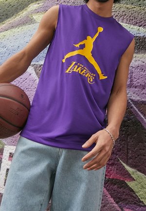 Person iført en lilla ærmeløs Los Angeles Lakers trøje, der holder en basketball foran en væg dækket med graffiti.