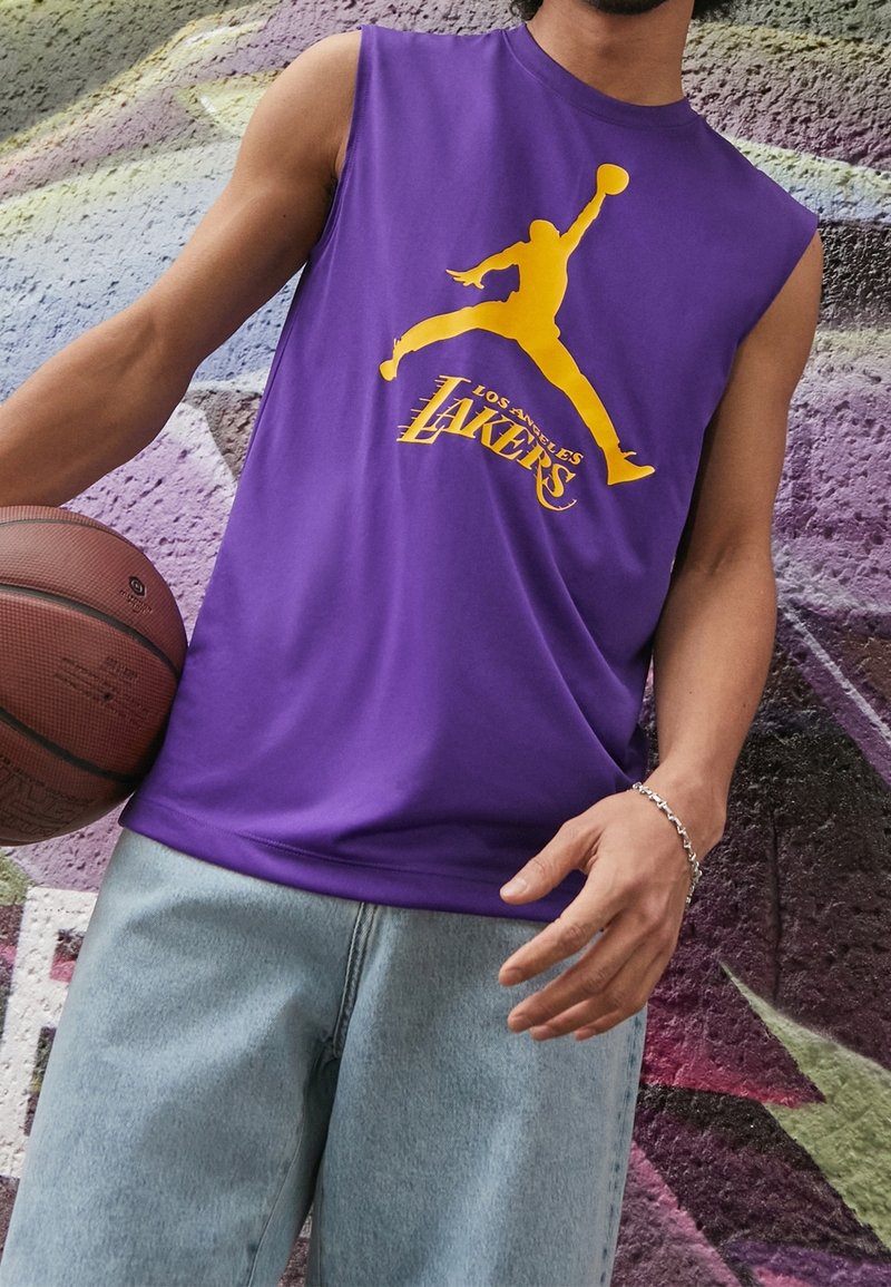 Personne portant un maillot sans manches violet des Los Angeles Lakers tenant un ballon de basket devant un mur couvert de graffitis.