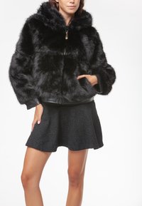 Giacca nera in pelliccia sintetica con cappuccio, caratterizzata da una chiusura a zip e finiture in pelle. La modella indossa una gonna nera con texture, che mette in mostra trame sovrapposte.