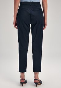 Pantaloni tapered blu navy con una texture liscia, dotati di due tasche posteriori e orlo arrotolato, abbinati a sandali neri con tacco.