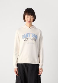 Person with short dark hair wears a cream hoodie with "GANT 1949 NEW HAVEN" text and black cargo pants, standing against a white background.