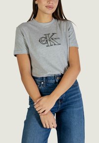 Szary bawełniany T-shirt z krótkimi rękawami, z czarnym nadrukowanym logo „Calvin Klein” oraz dużą literą „K” na środku klatki piersiowej.