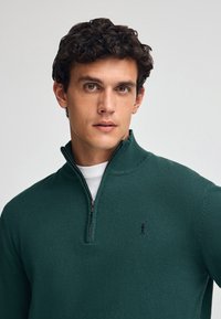 Grön zip-up tröja i strukturerad stickad kvalitet, med hög krage och en liten broderad logotyp på bröstet.