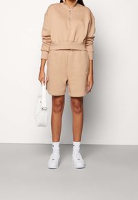 Beige Nike sweatshirt with half-zip, paired with beige shorts. White sneakers and a small white bag. Smooth fabric, casual fit.