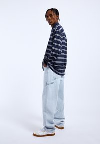 Camisa de rayas azul marino de manga larga con líneas blancas, combinada con jeans holgados de un azul claro. Zapatillas blancas con suelas marrones y sutiles acentos de marca.