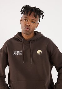 Brown hoodie made of soft fabric, featuring "THE CODE OF COOL" text and a pixelated yin-yang symbol on the front. Drawstring hood.