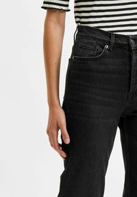 Svarta denimjeans med hög midja och fem fickor, med en subtil textur och en åtsittande silhuett. Randig skjorta synlig högst upp.