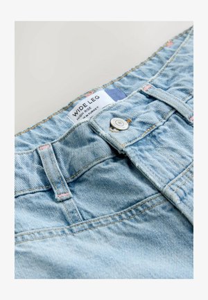 Heledad sinine laiapõhjaline denim püksid, millel on kõrge vöökoht, hõbedane nööbilukk ja kontrastne kollane õmblus. Silt on nähtav.