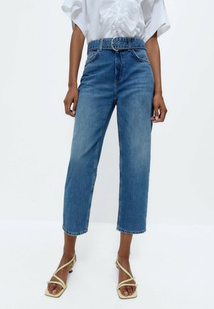 Femme portant un jean bleu taille haute avec une ceinture large et une coupe larges raccourcie, associé à des talons beiges à lanières et à bout carré, et un haut blanc.