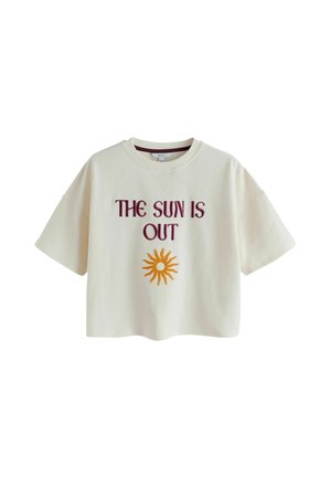 Kvinde med krøllet hår står på en klippefyldt strand iført en hvid skjorte med teksten "THE SUN IS OUT" og en sol-grafik, kombineret med leopardprint shorts.