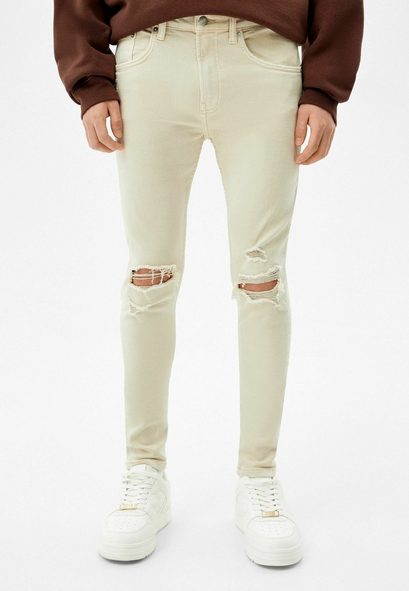 Jeans skinny desgastados color beige con varias áreas rasgadas en las rodillas. Combinados con zapatillas blancas que tienen un acento dorado y cordones texturizados.