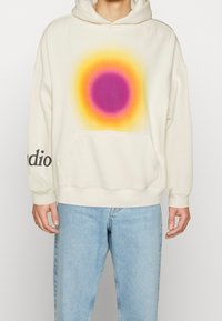 Lichte crème capuchontrui met een geleidelijke cirkelvormige print in roze en oranje, met een voorzak en de tekst "studio" langs de mouw.