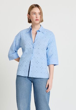 Femme aux cheveux blonds courts portant une chemise boutonnée bleu clair en tissu œillet et un jean bleu, debout devant un fond clair uni.