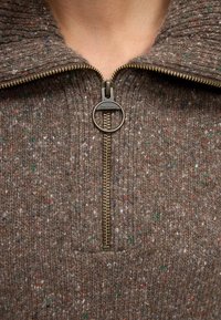 Gros plan d'un pull en laine marron tacheté avec un curseur de fermeture éclair circulaire en laiton Barbour partiellement ouvert au niveau du cou.