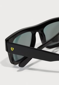 Sorte solbriller med grønne linser, der har en tykk ramme og Ferrari-logo på siden. Glat overflade og vinkelret design for moderne appel.