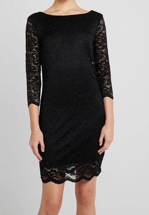 Robe en dentelle noire avec des manches trois-quarts transparentes et un ourlet festonné portée par une personne debout devant un fond uni.