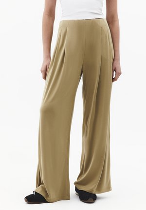 Femme portant un pantalon large beige ample avec des plis, associé à des baskets noires et un haut blanc, debout devant un fond blanc.