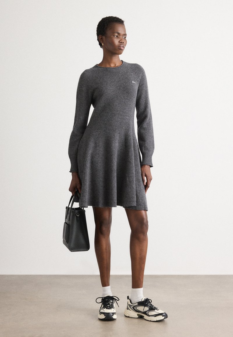 Abito grigio a maglia con maniche lunghe e gonna svasata, caratterizzato da un collo rotondo e un piccolo logo. Abbinato a sneakers nere e una borsa.