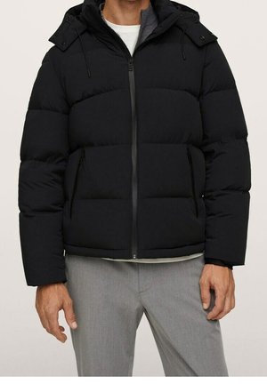 Homme portant une veste à capuche matelassée noire, zippée, avec un pantalon gris et une chemise blanche en dessous, visible du cou aux hanches.