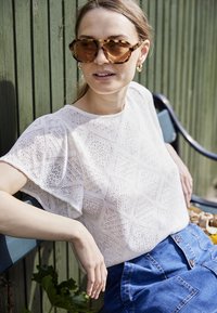 Sieviete ar gaiši brūniem matiem, kas valkā bruņu bruņu saulesbrilles, baltu adītu topu un zilu džinsa svārku, sēž zaļā sola ārā.