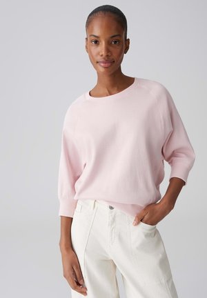 Femme aux cheveux courts portant un haut à manches longues rose clair et un pantalon taille haute blanc, debout avec une main dans la poche contre un fond uni.