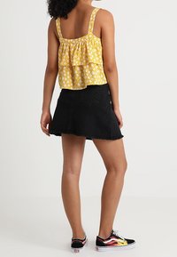 Top senza maniche con motivo floreale giallo e strati arricciati, abbinato a una gonna in denim nera e sneakers nere con dettagli a fiamma.