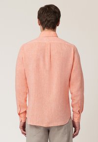 Chemise en lin corail clair avec des manches longues, un col boutonné et une texture subtile. Le dos possède un pli central et des poignets boutonnés.