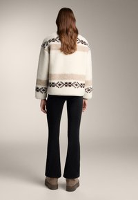 Kurtka z polaru z wzorzystymi detalami w beżu i brązie, w zestawie z czarnymi rozszerzanymi spodniami i beżowymi butami na platformie; teksturowana, o luźnym kroju.