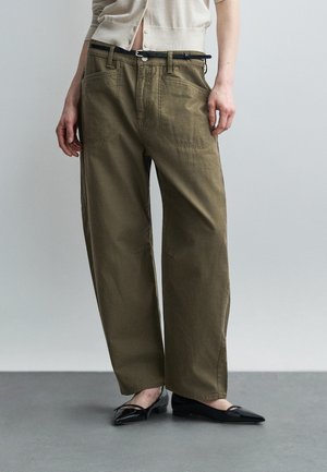 Person wearing loose olive green pants with large front pockets, black belt, beige top, and black pointed flats, standing on gray floor.