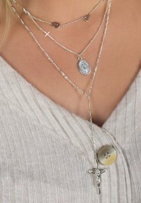 Silberne Schichtenketten mit einem Kreuz, ovalem Anhänger und dekorativen Gliedern auf einem strukturierten, hellbeigen Pullover.