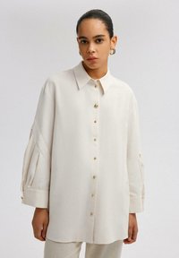 Camicia a maniche lunghe color crema con frontale a bottoni, colletto strutturato e maniche plissettate. Presenta bottoni in tono dorato e una vestibilità comoda.