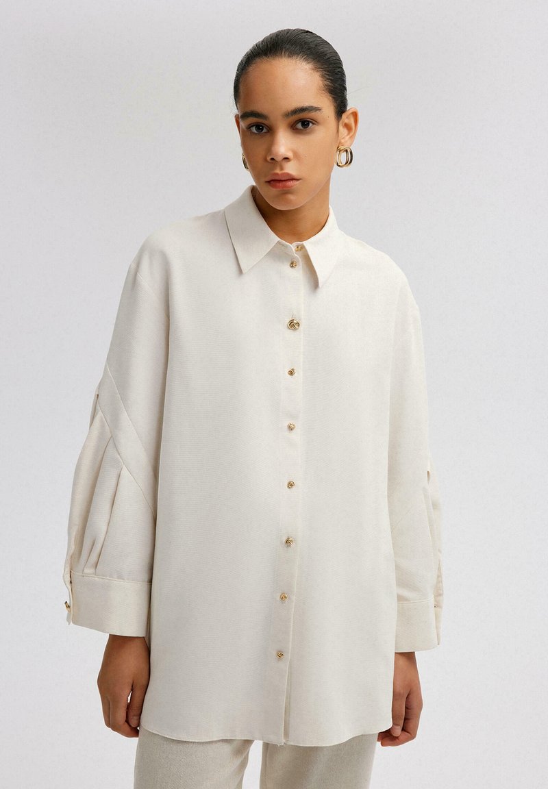 Camicia a maniche lunghe color crema con frontale a bottoni, colletto strutturato e maniche plissettate. Presenta bottoni in tono dorato e una vestibilità comoda.