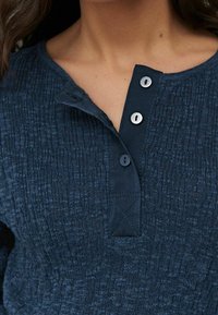 Haut en tissu texturé bleu foncé avec une patte de boutonnage comportant trois boutons gris et un col renforcé.