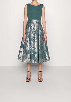 Robe sans manches couleur sarcelle avec une jupe ample à motifs floraux métalliques argentés, portée avec des sandales à talons hauts noires à brides, sur un fond uni.