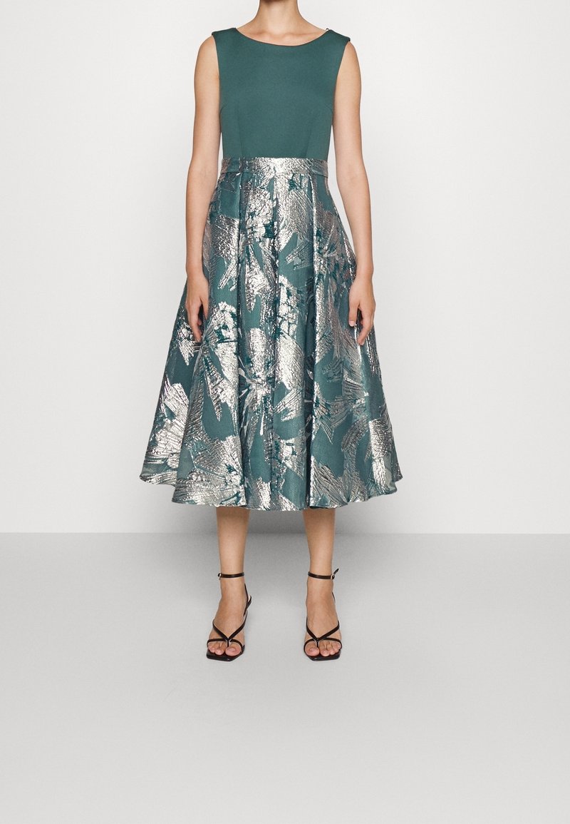 Robe sans manches couleur sarcelle avec une jupe ample à motifs floraux métalliques argentés, portée avec des sandales à talons hauts noires à brides, sur un fond uni.