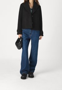 Černá zkrácená bunda se širokým límcem a dvěma knoflíky, ve spojení s modrými volně střiženými džíny a černými botami, drží černou tašku.