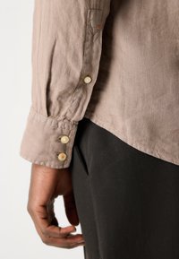 Camicia button-up marrone chiaro con una finitura texturizzata, dotata di tre bottoni ai polsini e una vestibilità comoda, abbinata a pantaloni neri.