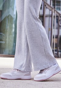 Des chaussures de sport blanches et gris clair avec des accents roses, un design texturé, des matériaux doux et un profil bas. Portées avec un pantalon côtelé gris clair.