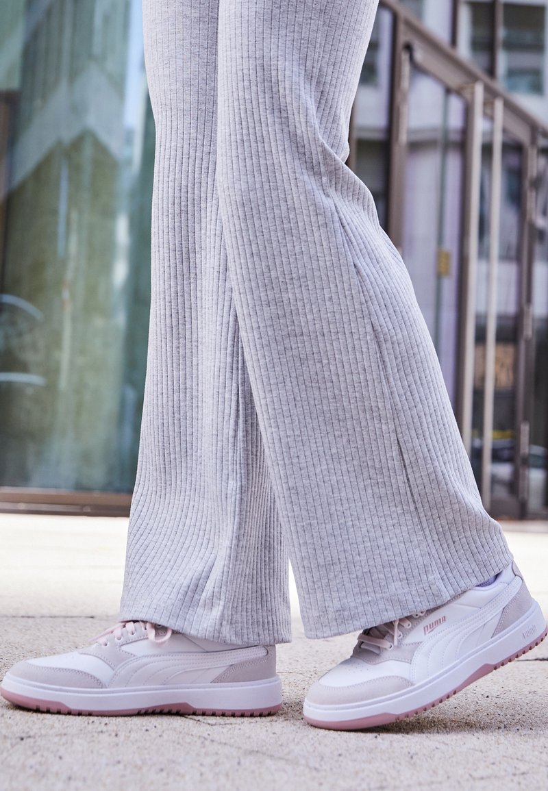 Des chaussures de sport blanches et gris clair avec des accents roses, un design texturé, des matériaux doux et un profil bas. Portées avec un pantalon côtelé gris clair.