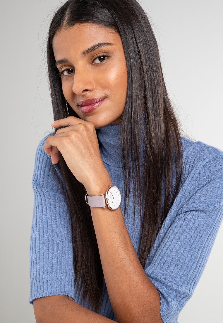 Eine roségoldene Uhr mit rundem Zifferblatt und hell-lila Armband, am Handgelenk eines Models getragen, kombiniert mit einem gerippten blauen Pullover.